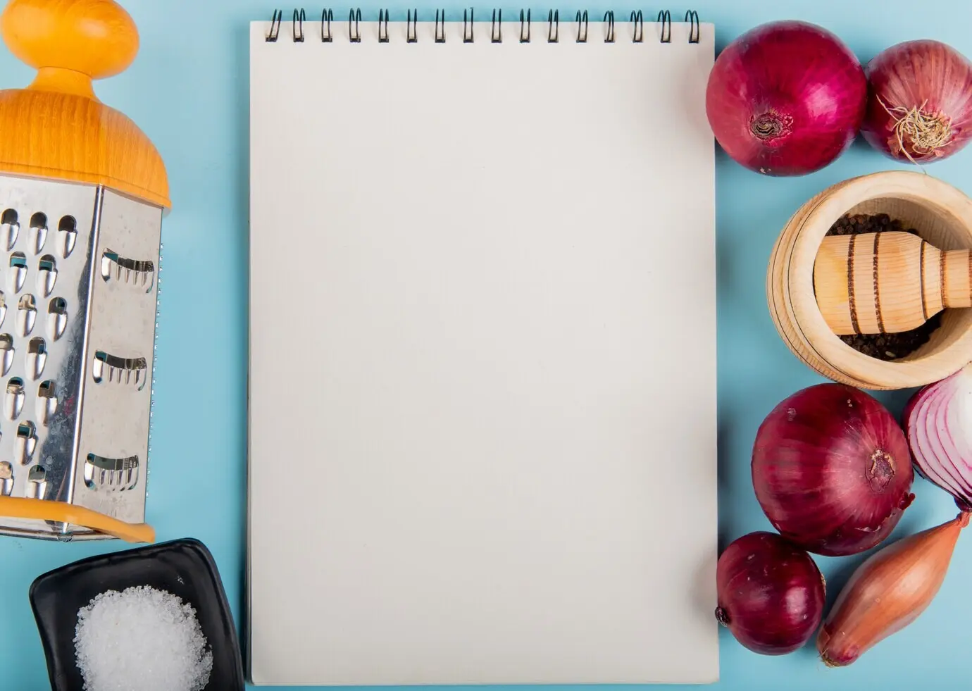 Draufsicht auf Zwiebeln mit Salz, schwarzen Pfefferkörnern und einer Reibe, um einen Notizblock herum, auf blauer Oberfläche mit Copyspace.