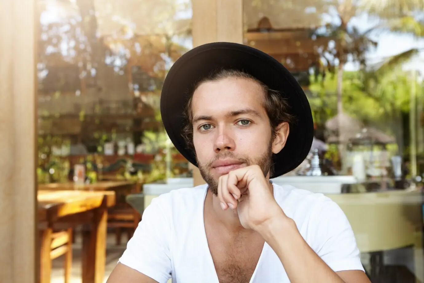 Kopfporträt eines modischen, attraktiven jungen Studenten mit stylischem Bart, der einen schwarzen Hut und ein Shirt mit V-Ausschnitt trägt und mit einem Lächeln blickt.