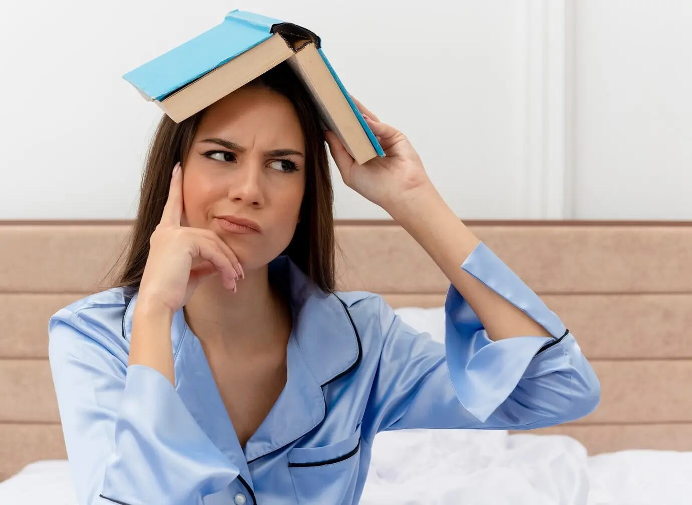 In einem Schlafzimmer vor hellem Hintergrund sitzt eine junge, schöne Frau im blauen Pyjama auf dem Bett, hält ein Buch über ihrem Kopf und blickt mit skeptischem Ausdruck zur Seite.
